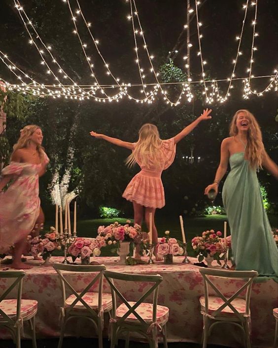 three women gleefully celebrate Midsummer by dancing on a table outdoors under fairy lights