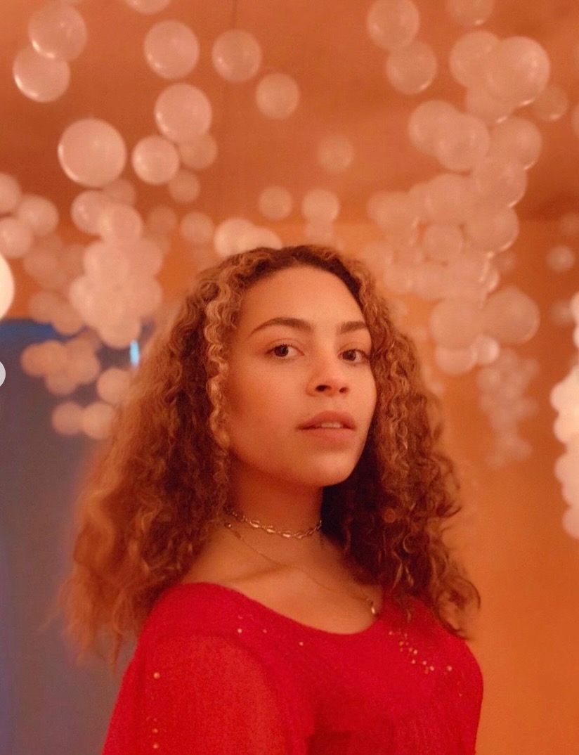 Photo of Shelby Edlund in reddish pink room surrounding by bubbles.