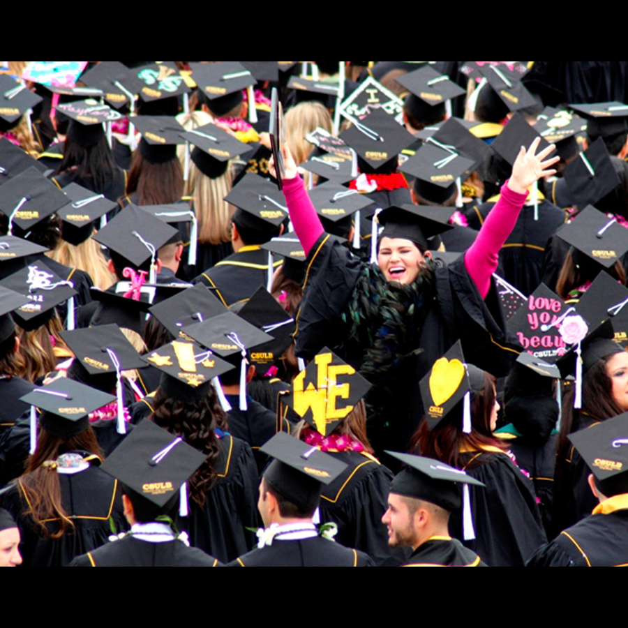 A college grad stands up on her chair to turn around and wave to her audience while the rest of the graduates are seated facing forward.