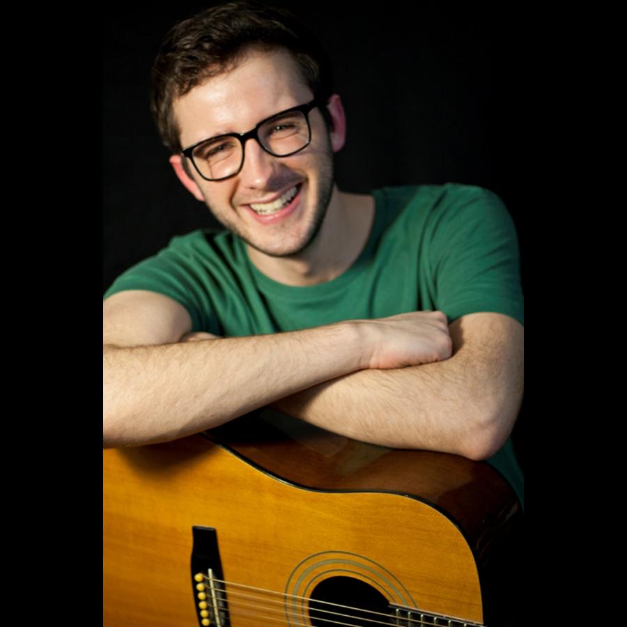 Man in glasses with his arms corssed leaning on his guitar.