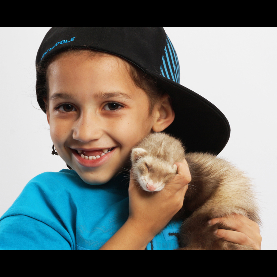 A close up of s little boy in a hat holding his pet ferret.