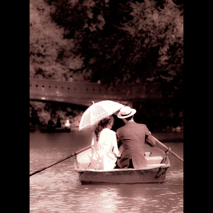 Bride and groom sitting next to each other sharing a parasol and on a boat the lake in Central Park.